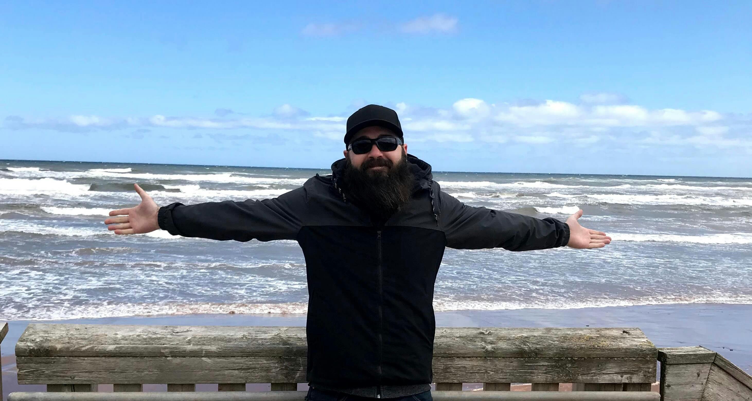 Background image of Dave with his arms out wide standing in front of the ocean